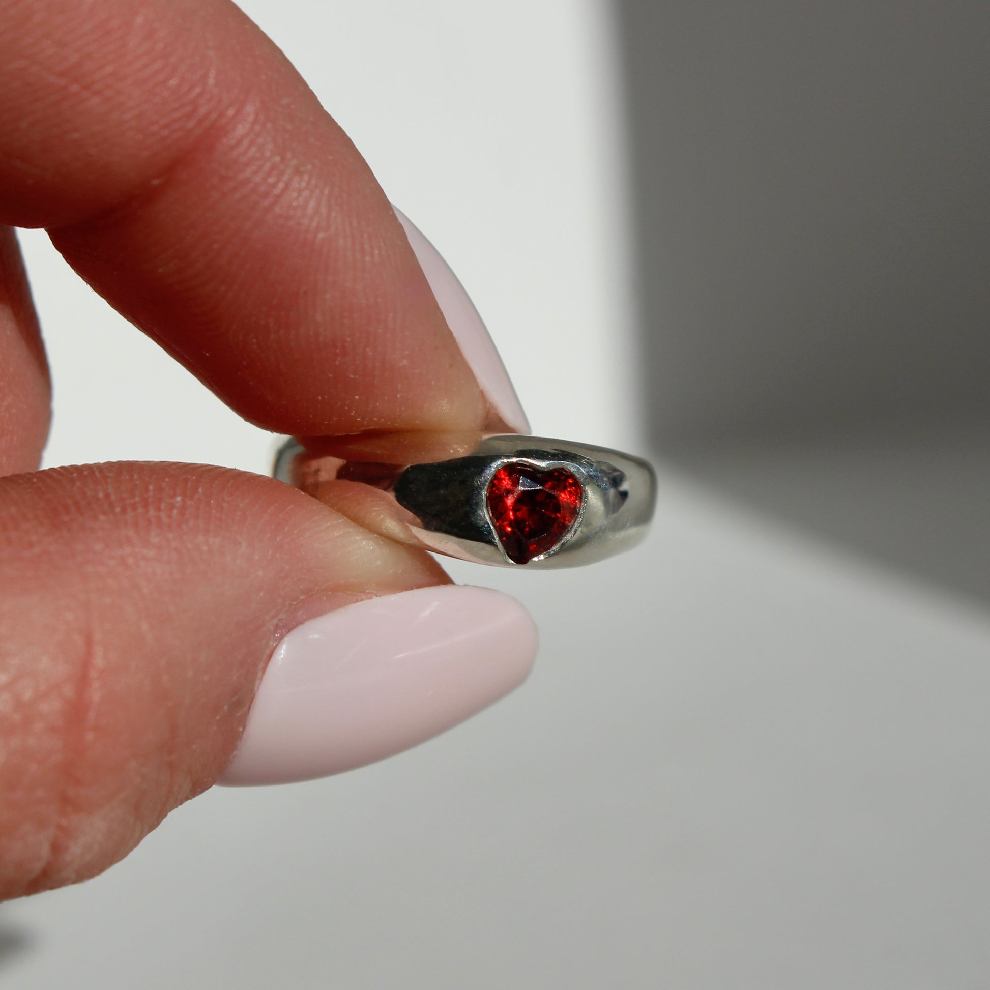 Silver ring with a red heart-shaped gemstone held between fingers against a neutral background